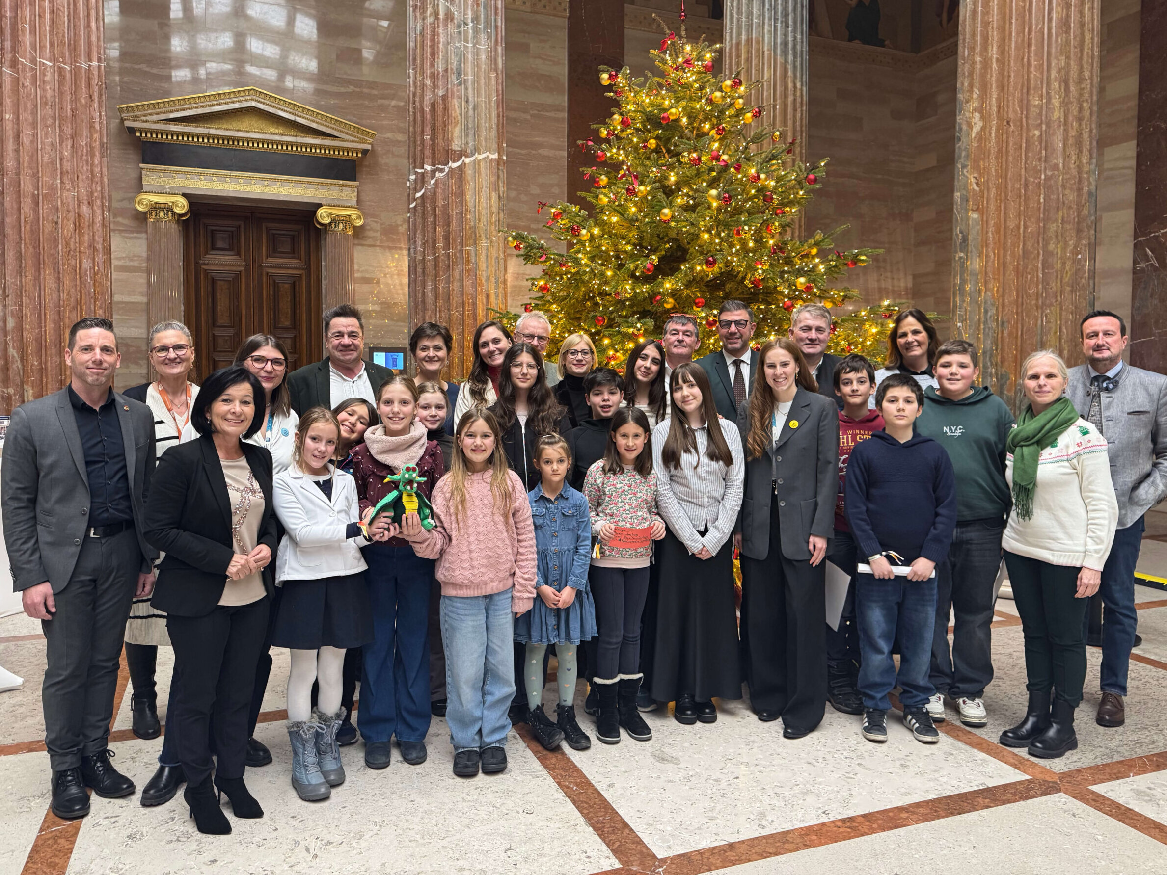 Gruppenfoto mit Kindern und Politiker*innen vor einem großen Weihnachtsbaum in der Säulenhalle des Parlaments.
