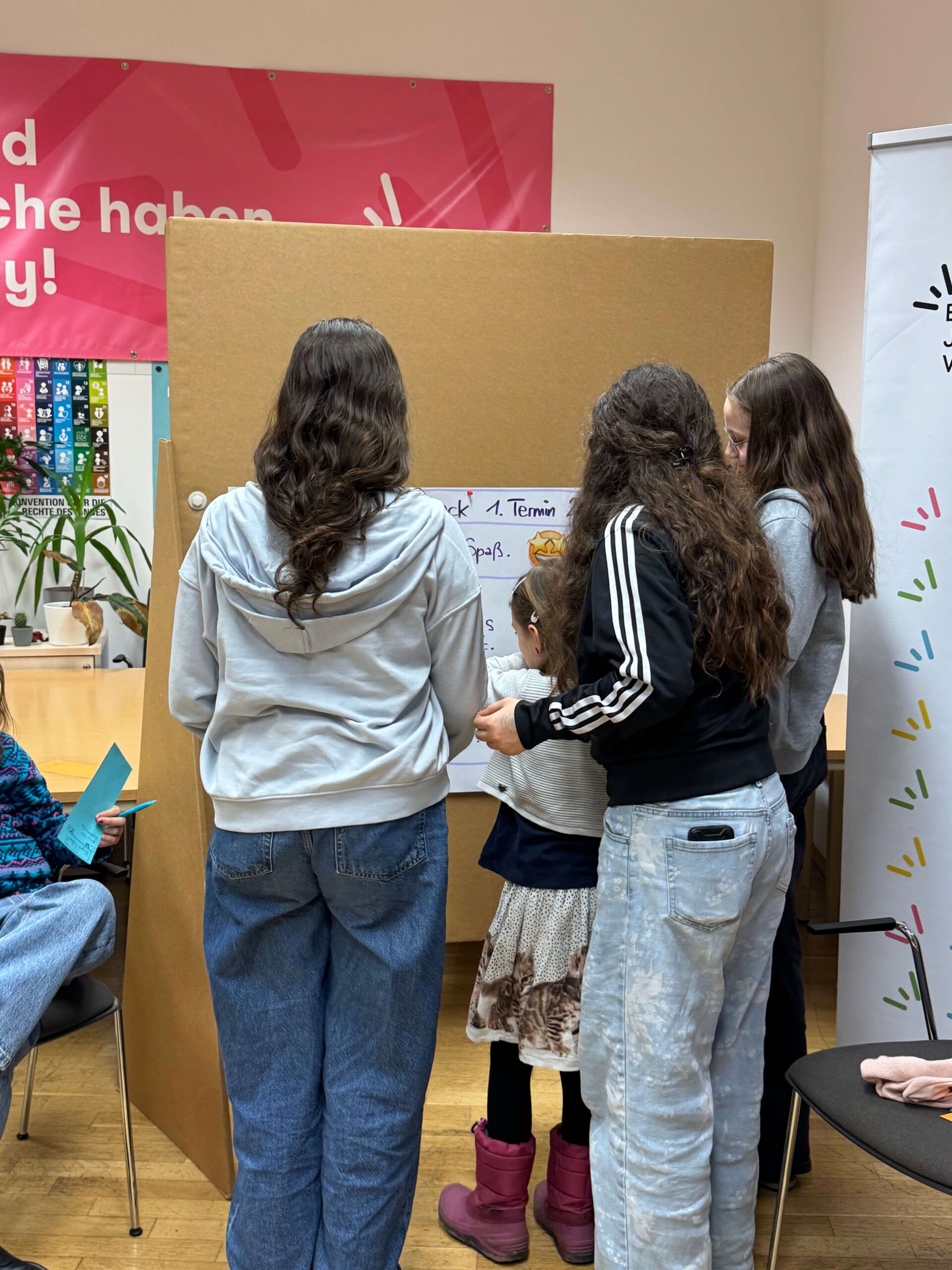 Vier Mädchen sind von hinten zu sehen, wie sie Sticker auf eine Pinnwand kleben. Im Hintergrund der Pinnwand hängen bunte Plakate.