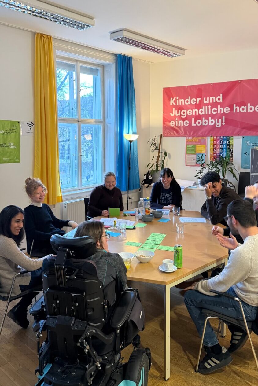8 Mitglieder des Inklusionsbeirats sitzen in einem Tischkreis und lachen.