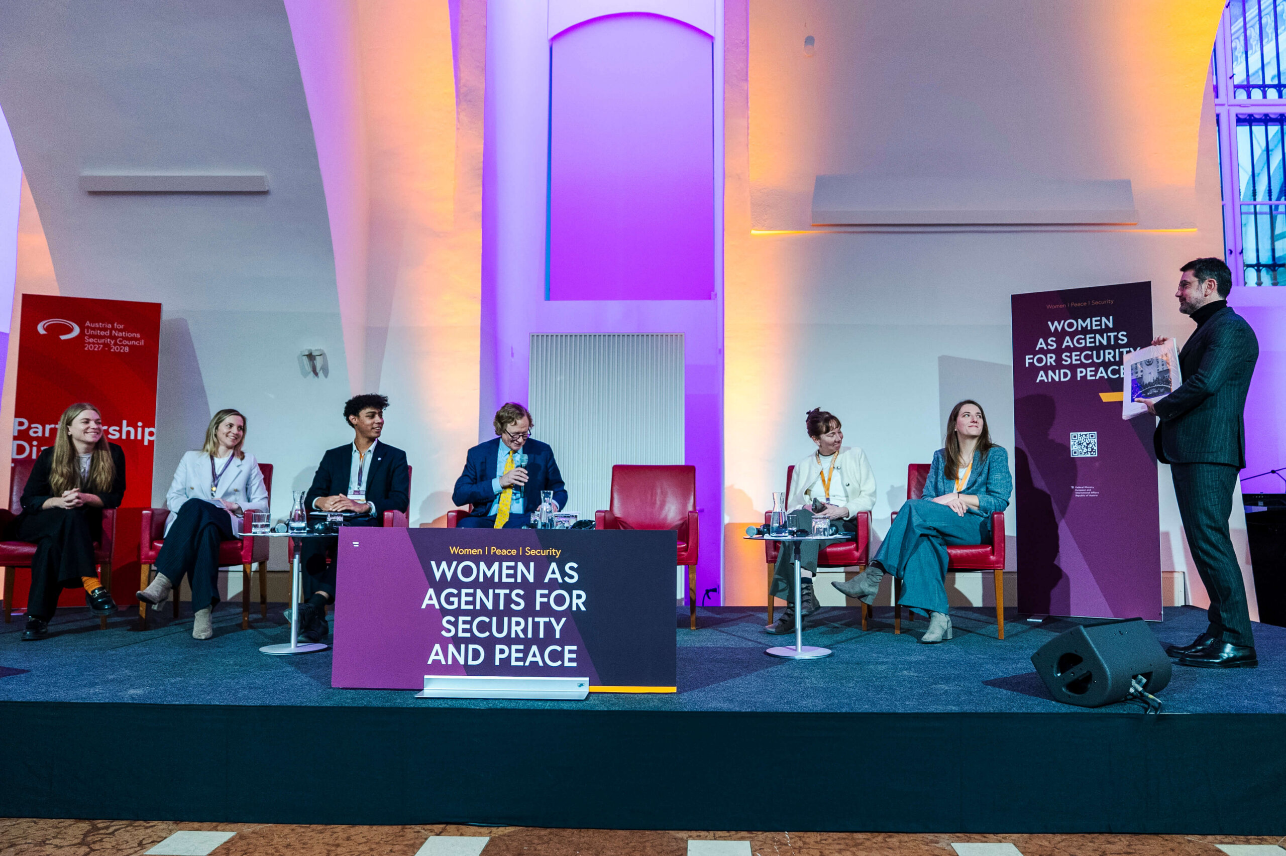 Sechs Personen sitzen und eine Person steht auf einem Podium. Vor ihnen steht ein Banner mit der Aufschrift "Women as agents for security and peace".