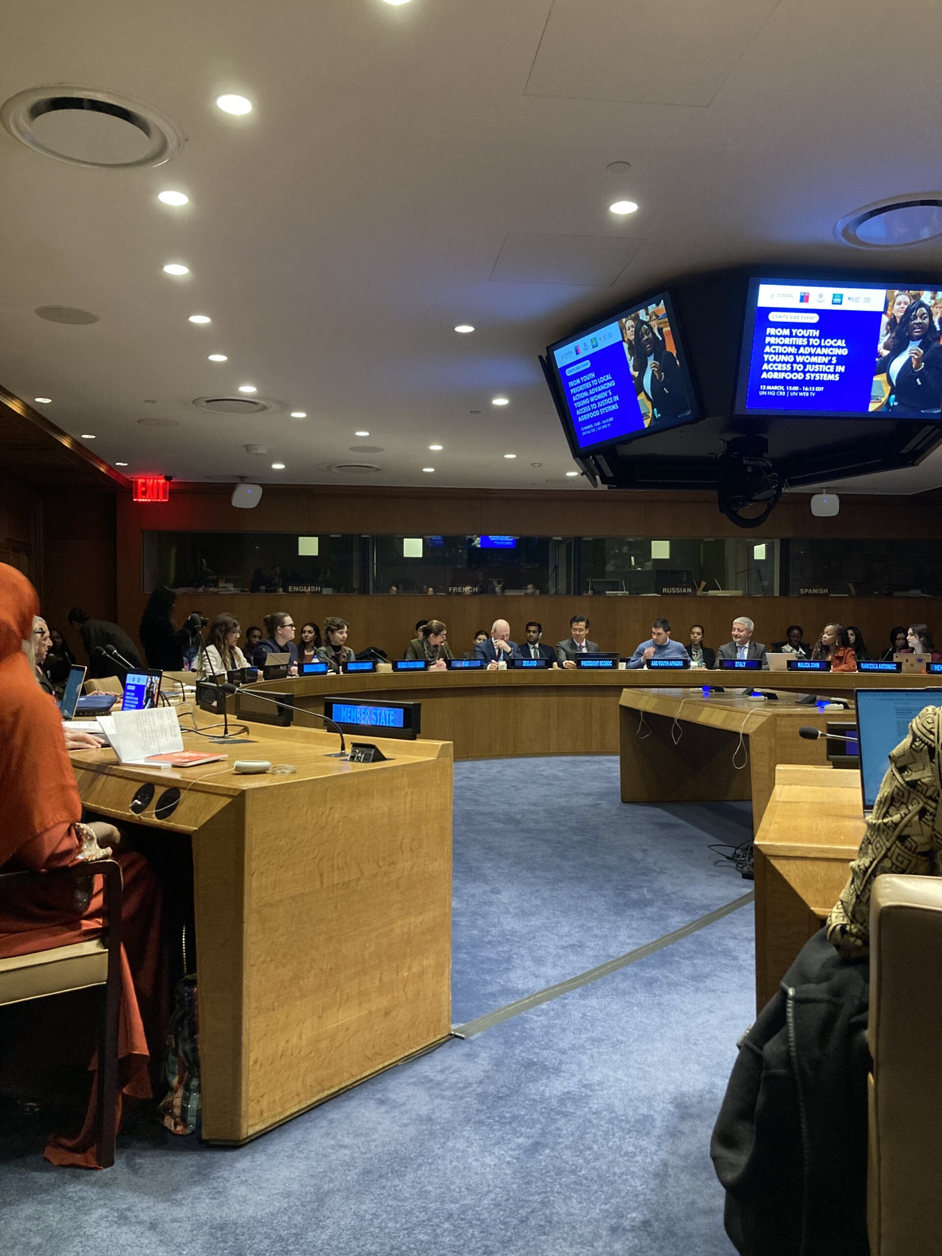 Ein runder Sitzungstisch mit verschiedenen Menschen. Ihr Herkunftsland wird auf digitalen Schildern eingeblendet. Auf einem Bildschirm in der Mitte wird der Titel "From Youth Priorities to local action: adcancing young women's access to justice in agrifood systems" eingeblendet.