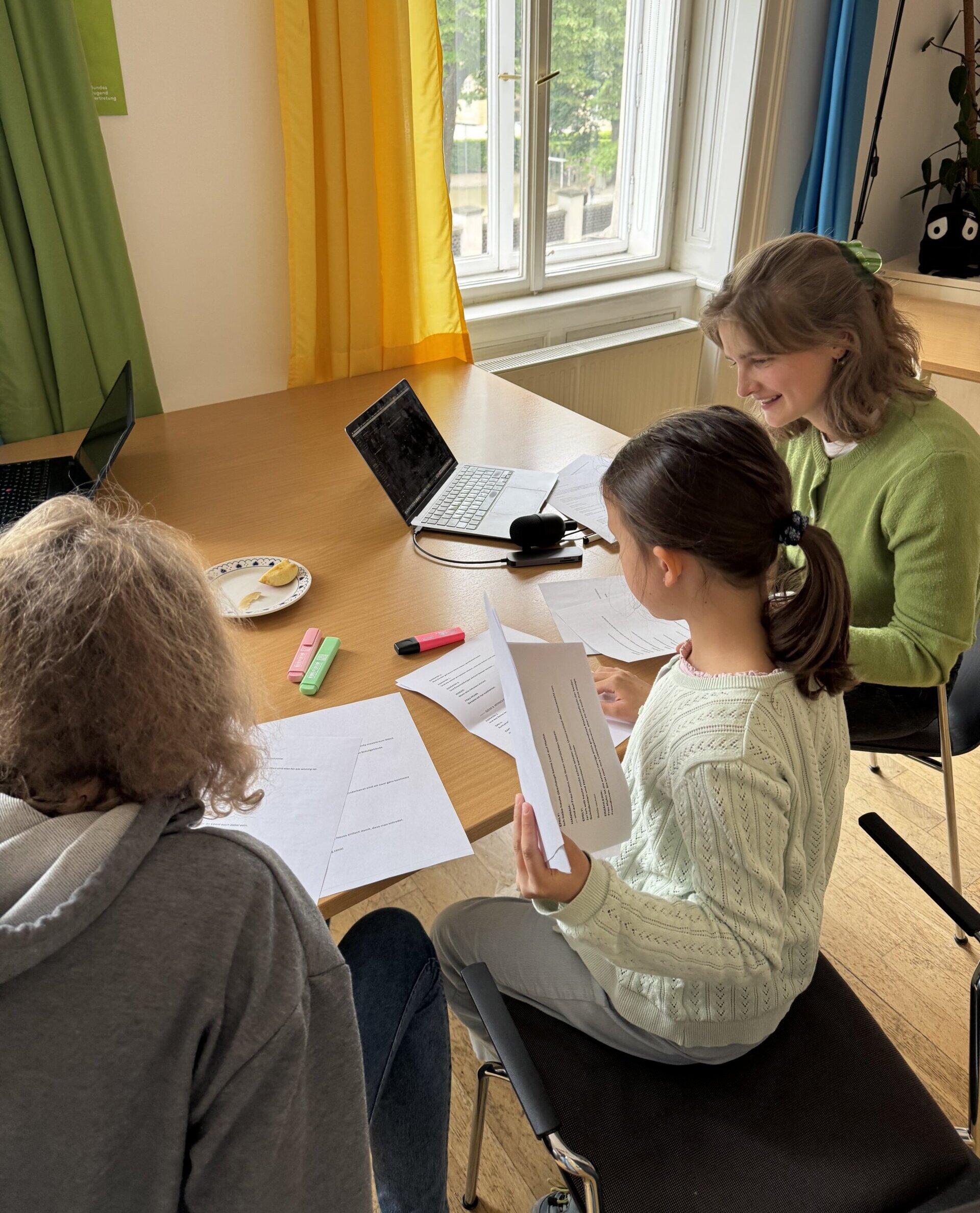 Kinderbeirat-Medienwerkstatt Zwei Mädchen und eine junge Frau sitzen an einem Tisch und lesen Skripte vor.
