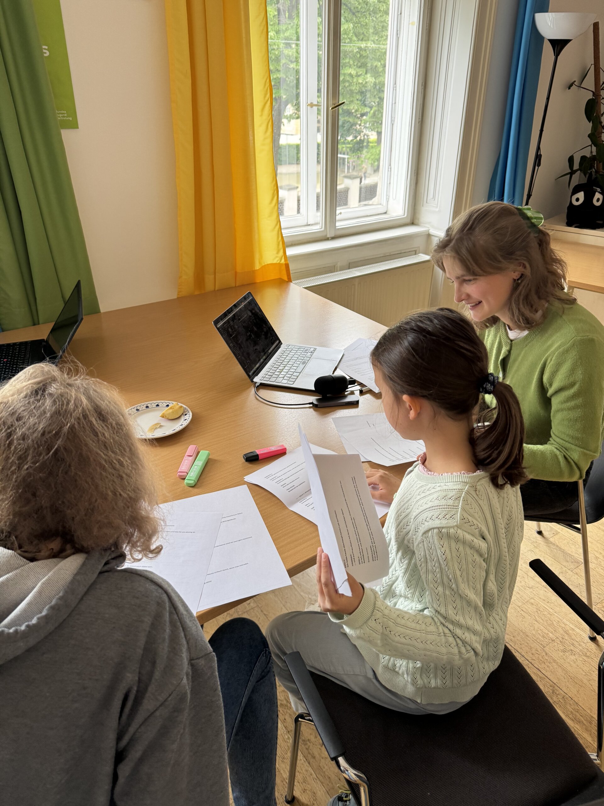 Zwei Mädchen und eine junge Frau sitzen an einem Tisch und lesen Skripte vor.